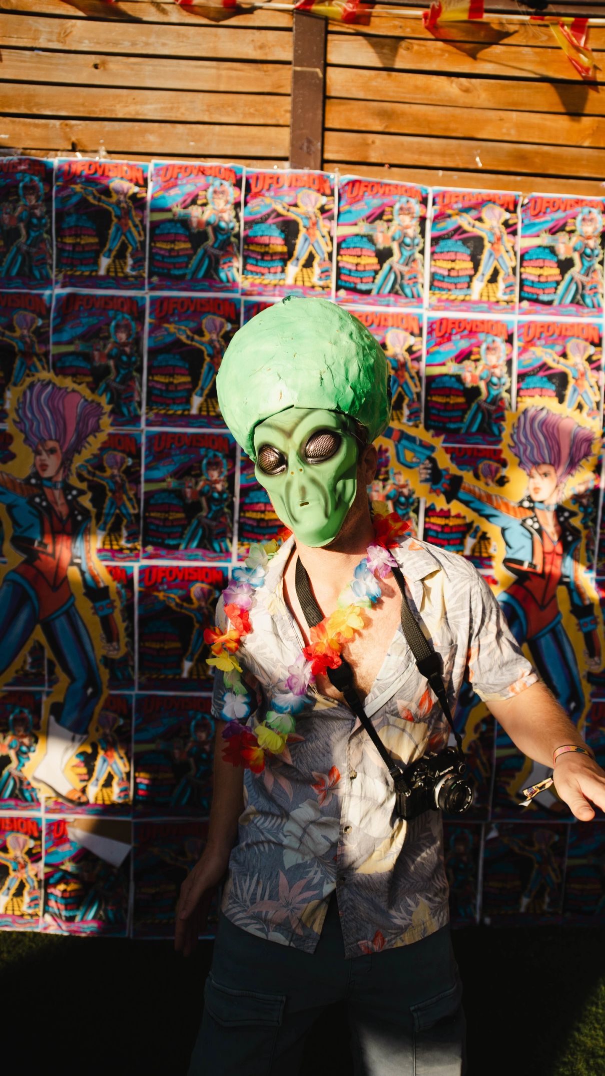 Yo con máscara de alien verde, collar de flores, camisa hawaiana y la cámara analógica al cuello, delante de una pared de carteles del Ufovision